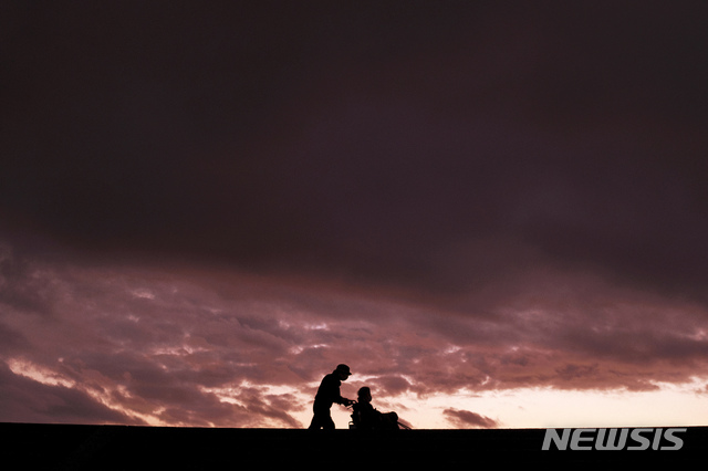 [도쿄=AP/뉴시스]지난 2020년 11월22일 일본 도쿄 남서부 시즈오카에서 마스크를 쓴 한 남성이 석양 하늘을 배경으로 방파제를 거닐면서 휠체어 탄 여성을 밀고 있다. 2020.11.23.