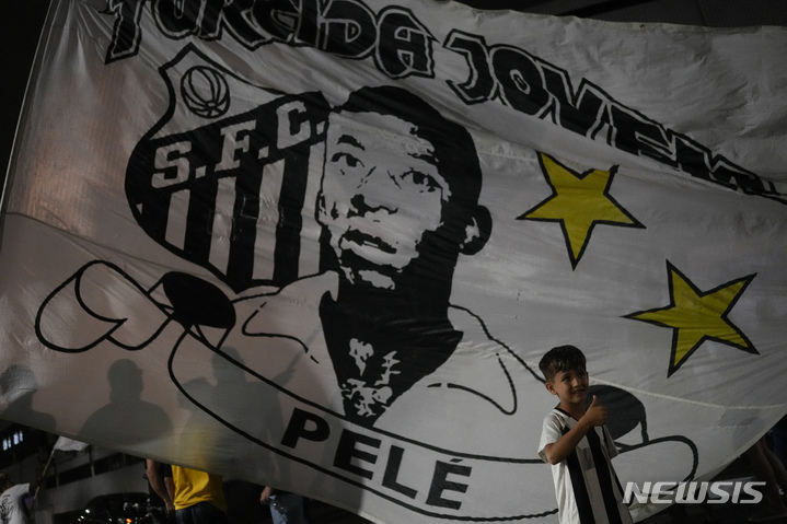[산투스( 브라질)= AP/뉴시스] 펠레의 죽음이 보도된 29일(현지시간) 브라질 산투스 축구장 앞에서 휘날리는 펠레 초상 깃발 앞에서 한 소년이 엄지를 들어보이고 있다.  