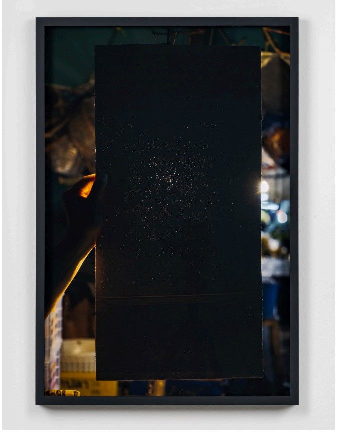 프랏차야 핀통, 〈"."〉, 2018 피그먼트 프린트, 액자, 68.3 x 45.5 cm (액자 71.6 x 48.7 x 3.4 cm. 밤하늘인 듯 했던 평면이 누군가의 손에 들려 있다. 이건 사실 복권을 꽂아 팔고 난 뒤 그것이 꽂혀 있던 흔적만 남은 골판지 판이다. 대박을 꿈꾸며 복권을 산 사람의 흔적이 구멍으로 남은 것이다. 골판지를 들고 있는 사람은 툭툭버스 운전사나 라비올리 가판대 주인과 같은 길거리 상인들이다. 골판지 판 너머로 각 상인의 가판대나 차에서 비추는 불빛이 구멍으로 이뤄진 별자리를 더욱 선명하고 아름답게 수놓는다. 주변의 현실과는 동떨어진 듯한 그 모습은 애초에 골판지에 구멍을 내게 한 덧없는 희망을 보여주는 듯 하다. 각각의 구멍은 단꿈에 젖어 복권을 산 사람 하나하나를 상징하고, 일확천금의 꿈으로 그 모두를 유혹한 경제적 추상화의 구조 그 자체와 다를 바 없어 보인다. *재판매 및 DB 금지