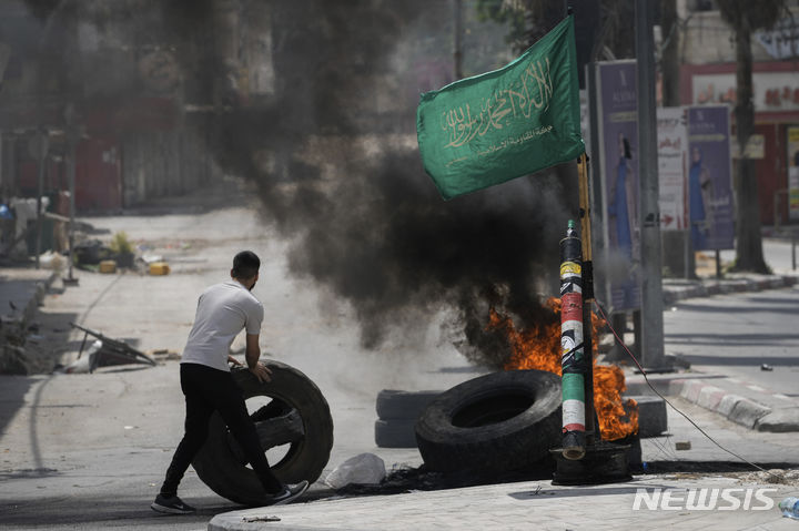 [제닌=AP/뉴시스] 22일(현지시각) 요르단강 서안지구 제닌에서 팔레스타인 시위대와 이스라엘군이 충돌한 가운데 한 남성이 하마스 깃발 옆에서 타이어를 태우고 있다. 2024.05.23.
