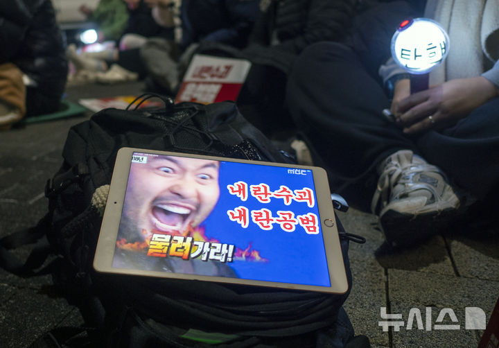 [전주=뉴시스] 강경호 기자 = 13일 전북 전주시 전주객사 풍패지관 앞에서 열린 비상계엄 선포 규탄 및 정권 퇴진 촛불집회에 참석한 한 시민이 예능프로그램인 '무한도전'의 장면에 글귀를 합성한 사진을 태블릿에 띄워놓고 있다. 2024.12.13. lukekang@newsis.com