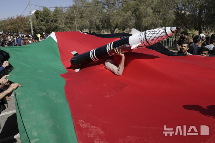 [사나=AP/뉴시스] 올 해 1월 22일 예멘 사나에서 반미·반이스라엘 시위가 열려 대형 팔레스타인 깃발 틈으로 얼굴을 내민 한 후티 반군 지지자가 모형 로켓을 들고 시위를 벌이고 있다. 2025.03.05.