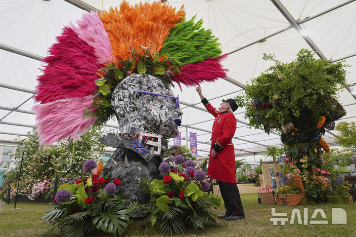 Chelsea Pensioner Peter Wilson reaches up to touch a Punks head with a mohawk created with pampas grass, tropical blooms and preserved leaves part of Chelsea in Bloom by Ricky Paul Flowers at the RHS Chelsea Flower Show in London, Monday, May 19, 2025. (AP Photo/Kirsty Wigglesworth)