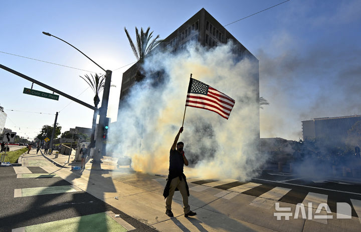 [산타애나=AP/뉴시스] 9일(현지 시간) 미 캘리포니아주 로스앤젤레스 남부 산타애나 연방 청사 앞에서 한 시위자가 경찰이 쏜 최루 가스 속에서 성조기를 흔들고 있다. 2025.06.10.