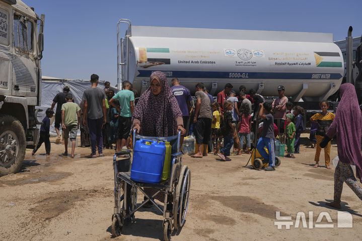 [AP/뉴시스] 19일 가자 남부 칸유니스에서 급수 탱크차 앞에 사람들이 줄울 서 있고 한 여성이 휠체어로 플라스틱 물통을 밀어 나르고 있다