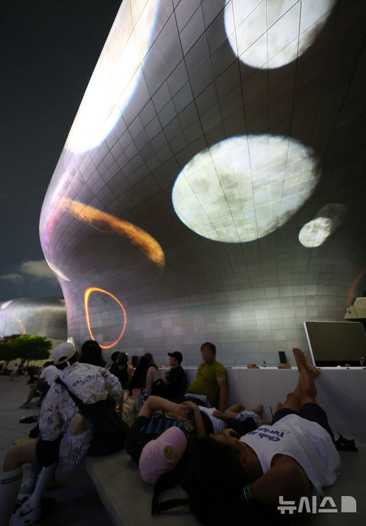 [서울=뉴시스] 조성우 기자 = 28일 오후 서울 중구 동대문디자인플라자(DDP)에서 열린 '서울라이트 DDP 2025 가을'을 찾은 시민들과 외국인 관광객들이 미디어파사드 작품을 관람하고 있다. 해당 미디어 파사드는 오픈AI 지원으로 영상 생성 플랫폼 '소라(Sora)'를 활용해 제작한 티모 헬거트의 '문 사이클'. 2025.08.28. xconfind@newsis.com