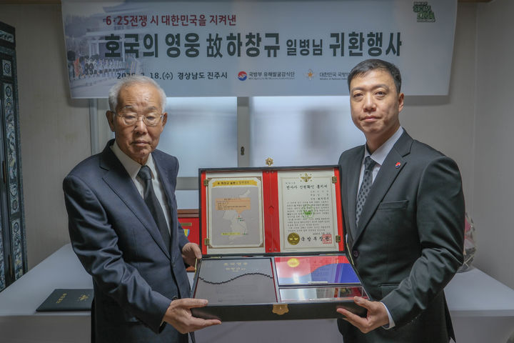 [서울=뉴시스] 18일 오전, 경남 진주시 소재 고 하창규 일병의 아들 하종복 씨(왼쪽) 자택에서 열린 호국영웅 귀환행사에서 김성환 국방부 유해발굴감식단장 직무대리(육군 중령, 오른쪽)가 전사자 신원확인 통지서 등이 담긴 호국의 얼 함을 하종복 씨에게 전달하고 있다. (사진=국방부 제공) 2026.03.18. photo@newsis.com *재판매 및 DB 금지