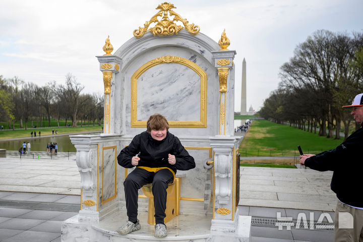 [워싱턴=AP/뉴시스] 30일(현지 시간) 미국 워싱턴DC 내셔널 몰에서 방문객이 ‘왕에게 어울리는 왕좌'(A throne fit for a king)라는 제목의 황금 변기에 앉아 기념 촬영을 하고 있다. 이 변기는 게릴라 예술그룹 '시크릿 핸드셰이크'라는 단체가 만든 것으로 백악관 리모델링에 집착하는 도널드 트럼프 대통령을 조롱하는 작품이다. 2026.03.31.