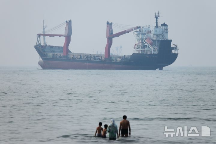 [반다르아바스=AP/뉴시스] 일본 정부가 나프타 비축 의무화를 다시 추진한다. 9일 마이니치신문 등 현지 언론에 따르면 일본 정부는 호르무즈 해협 불안으로 나프타 수급 차질이 커지자 30여 년 만에 비축 의무화 재도입을 검토하고 있다. 사진은 지난 6월30일(현지시간) 이란 반다르아바스 앞 호르무즈 해협에서 화물선 한 척이 정박해 있는 가운데 사람들이 얕은 물에 서 있는 모습. 2026.07.09.