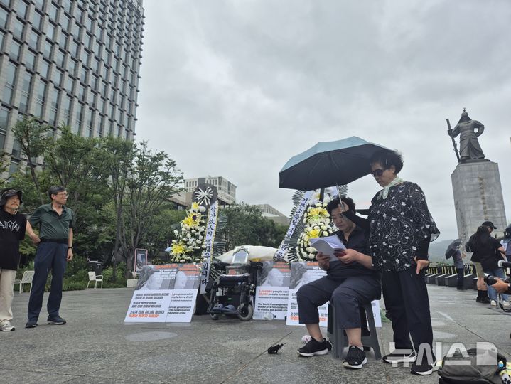 [서울=뉴시스] 조성하 기자 = 전국가습기살균제참사피해자연대가 17일 오전 서울 종로구 광화문광장 고(故) 서영철 대표의 영정 앞에서 우산을 쓴 채 기자회견문을 읽고 있다. 2026.08.17. create@newsis.com