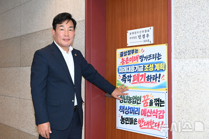 [창원=뉴시스] 경남도의회 농해양수산위원회 민경우 부위원장이 7일 도의회 3층 자신의 의원연구실 출입문에 부착한 정부의 미래대응기금 조성계획에 반대한다는 내용의 글귀 앞에서 포즈를 취하고 있다.(사진=민경우 도의원 제공) 2026.10.07. photo@newsis.com